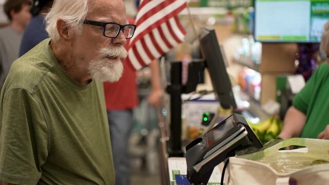 Elderly man enjoys his time at the grocery store checkout as he pays with a credit card after shopping for food and other items