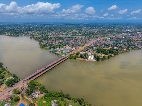 Aerial view of the Mesurado River bridge connecting urban areas in Monrovia, with dense buildings, lush vegetation, and a wide river under a blue sky. Monrovia, Montserrado, Liberia.