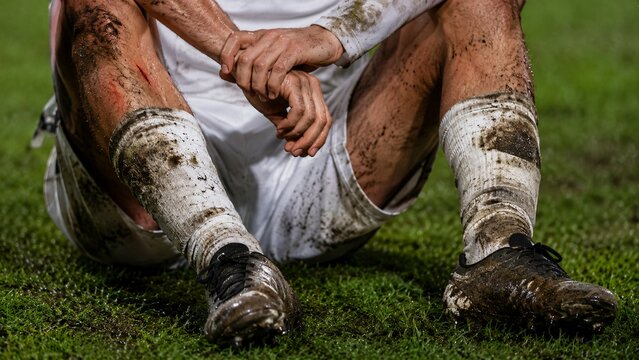 Exhausted muddy soccer player sitting defeated on the green grass pitch after a hard match