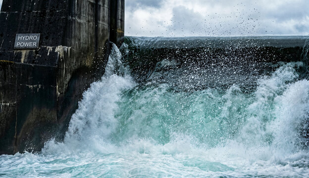 Powerful rushing water cascades over concrete dam edge at hydroelectric power plant generating clean renewable energy, water splash, blue and white foam.