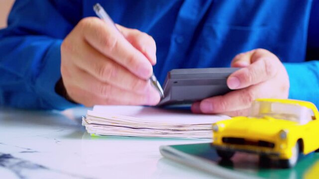 Businessman holding calculator and writing notes in notepad at desk. Focus on hands, yellow toy car on table. Close up, shallow depth of field, natural daylight. Financial audit  in autobusiness.