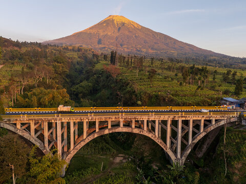 Aerial view of a concrete arch bridge spanning a lush valley with a majestic volcano in the background under golden hour light in Temanggung Regency, Central Java, Indonesia.