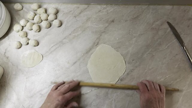 Overhead view of cook rolling thin dough circles with a wooden pin on kitchen counter