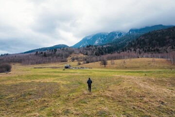 Obraz premium A lone traveler walks through a vast, peaceful meadow, framed by majestic mountains and a moody sky. This scene captures the essence of solitude, nature's beauty, and the journey of self-discovery