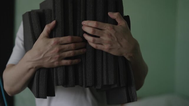 Cropped shot of man in white shirt carries stack of charcoal acoustic foam panels, preparing to soundproof room for recording studio, podcasting, music production. DIY home improvement project.