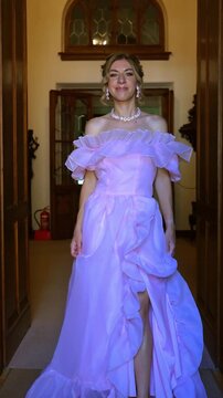 A beautiful confident mid-adult woman in a gorgeous pink ruffled gown looking at the camera and smiling, posing in a classic hall doorway. Vertical video.