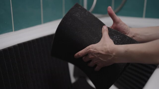 Person washing and wringing out black acoustic foam panels in bathtub. Squeezing water from soundproofing material for home studio or music room. DIY cleaning and preparation process.
