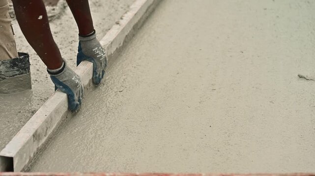 Builder using a straightedge tool to smooth and level a freshly poured concrete slab on a site