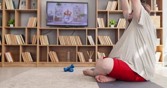 Overweight man practices yoga during online workout. He stretches on a mat at home, following a fitness video on TV, with breathing, balance, gentle exercise and meditation. Calm fit life at home.