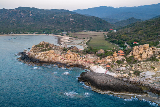 Aerial view of the rocky coastline at Bai Hom featuring rugged cliffs, a sandy beach, and lush green hills in Vinh Hai, Khanh Hoa, Vietnam.