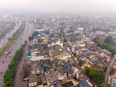 Aerial view of the dense urban landscape featuring residential rooftops with solar panels, a busy highway with traffic, and hazy atmospheric conditions in Lahore, Punjab, Pakistan.