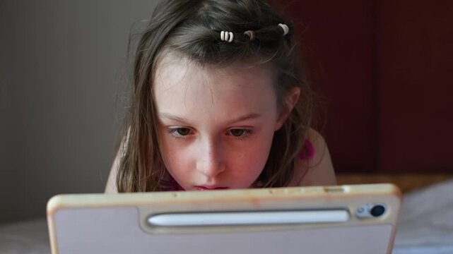 Focused girl watches videos on tablet while resting on bed, absorbed in digital entertainment during quiet leisure time.