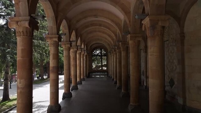 Walk through the beautiful stone colonnade of the historic Jermuk Mineral Water Gallery in Armenia, showcasing classical architecture with elegant arches and columns on a bright sunny day.

