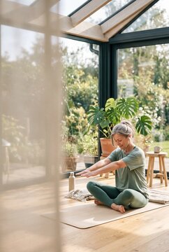Peaceful mature woman practicing yoga and meditation in a bright, modern sunroom surrounded by lush green plants
