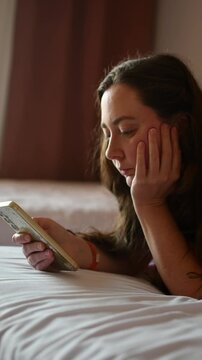 Young woman lies on hotel bed and scrolls smartphone with bored expression during quiet downtime alone in softly lit room.