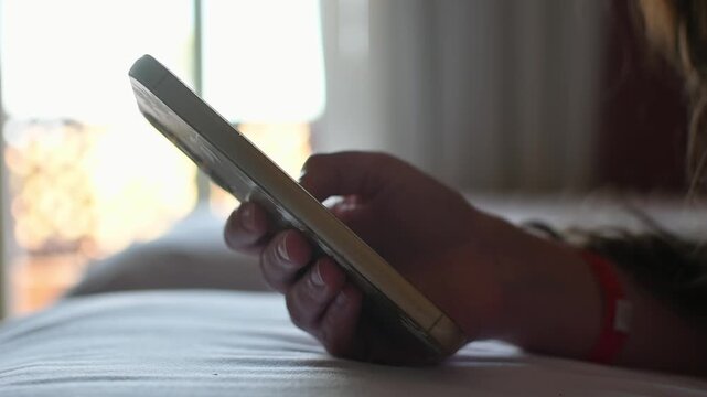 Young woman uses smartphone on hotel bed near window to stay connected and pass quiet leisure time during daytime indoors.
