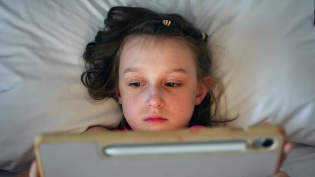 Young girl lies in bed and watches cartoons on tablet, enjoying quiet screen time and digital entertainment before sleep.