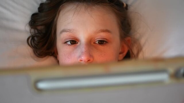 Close-up portrait of young girl lying in bed and watching cartoons on tablet during quiet screen time before sleep at home.
