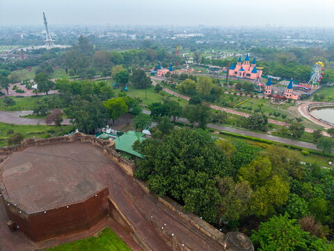 Aerial view of Minar-e-Pakistan and the surrounding Iqbal Park, featuring lush green lawns, pathways, and a distant amusement park with a Ferris wheel in Lahore, Punjab, Pakistan.