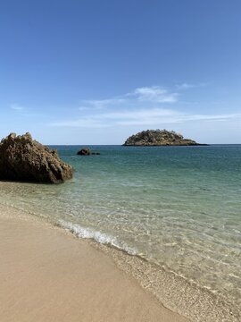 Praia de areia dourada com &aacute;guas cristalinas e ilh&eacute;u rochoso ao fundo sob c&eacute;u azul, paisagem costeira tranquila de ver&atilde;o.