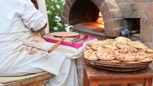Woman baker in headscarf places traditional flatbread into brick oven beside basket of fresh loaves at small outdoor bakery.