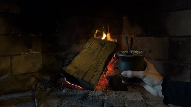 Close up of a hand holding traditional argentine yerba mate drink in front of a cozy burning fireplace a winter day