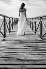 Fototapeta premium Woman in white dress walks on a pier while looking away into the distance at sunset