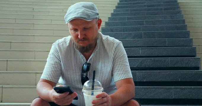 Man using smartphone and iced drink while sitting on stairs, wearing cap and striped shirt, with sunglasses hanging from collar in casual urban setting, relaxed pose and everyday city style.