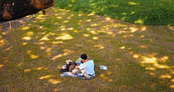 Overhead picnic on sunlit grass with couple seated on blanket, one person leaning over belongings, shoes nearby, framed by tree shade and open park lawn, relaxed outdoor scene with casual gathering