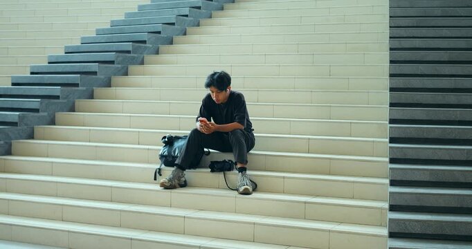 Young man sitting on stairs uses smartphone beside backpack in modern interior, isolated on wide cream steps with repeating geometric lines, quiet urban travel setting, relaxed waiting pose.