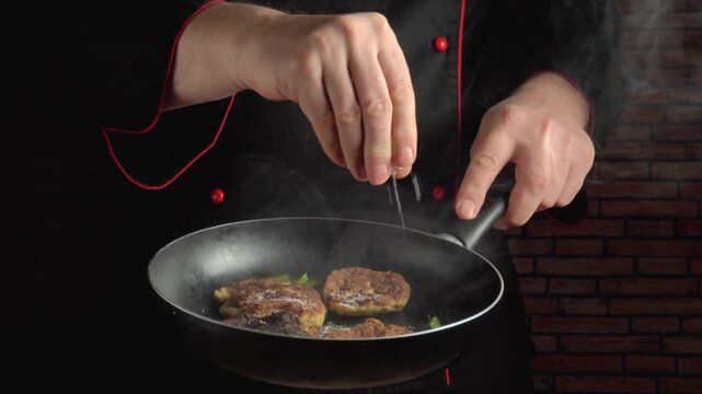 Chef in black uniform sprinkles seasoning over sizzling patties in a frying pan, with steam rising and a brick wall background enhancing the culinary scene