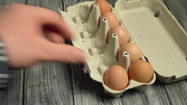 Hand reaches for brown eggs in a carton, removing one while others remain in the egg carton on a wooden surface
