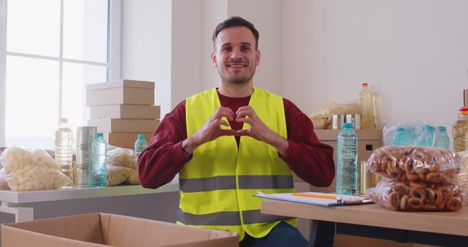 Smiling male volunteer standing in humanitarian aid center and making heart gesture with hands. Joyful young man supporting charity initiatives, helping food banks and social assistance programs.