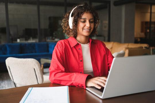 Young indian woman with curly hair and glasses working on laptop computer at desk in modern office or home workspace, wearing red shirt and smiling. Latin or muslim arab student girl studying indoors