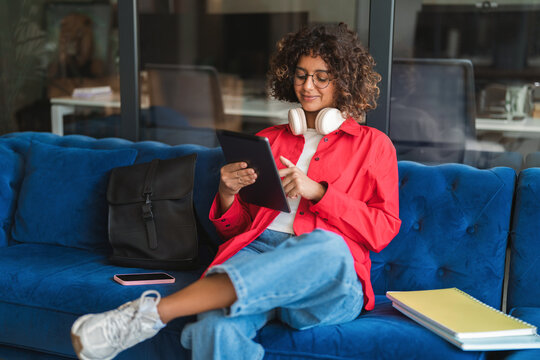 Young latin woman student on sofa using digital tablet technology for education, online browsing and studying in modern home office or campus. Freelance indian or muslim girl holding tablet for work