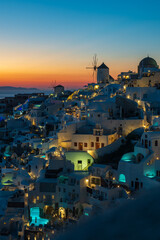 Twilight view of whitewashed houses and windmills glowing above the Aegean Sea in Oia, Santorini, Greece. © Andrei Tomas