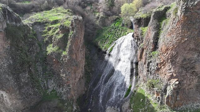 An aerial drone shot captures the majestic Jermuk Waterfall, known as Mermaid's Hair, cascading down rocky cliffs amidst lush green vegetation in Armenia's beautiful mountain landscape during spring.
