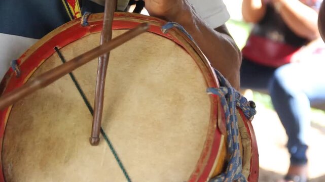 Hand holds drum with wooden sticks. Drum skin taut, red rim, green tension lines. Cultural rhythm, traditional music, celebration mood. Lamas, Tarapoto - Peru