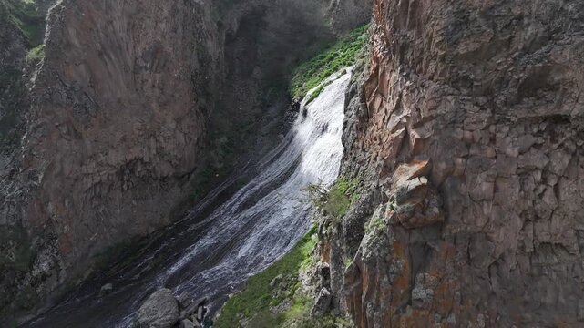 An aerial drone shot captures the majestic Jermuk Waterfall, known as Mermaid's Hair, cascading down rocky cliffs amidst lush green vegetation in Armenia's beautiful mountain landscape during spring.

