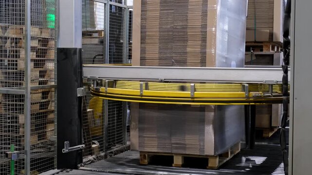 An automated factory packaging system wraps a tall stack of corrugated cardboard sheets on a wooden pallet using transparent plastic stretch film.