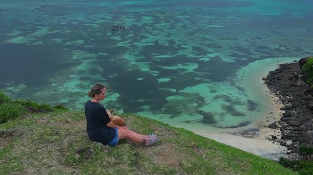 Caucasian woman sitting cliffside with dog overlooking turquoise ocean reef, traveler resting on grassy headland, panoramic aerial view of coral patches and sandy bay, Bukit Merese, Lombok