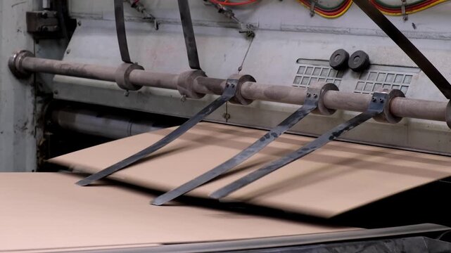 Metal tension guides and spring straps pressing down on flat cardboard sheets moving along a factory conveyor line.