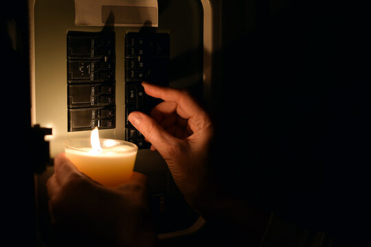 Close-up side view showing hand flipping circuit breaker switch in electrical panel while holding lit candle for illumination during blackout