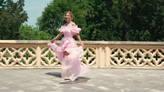 Cheerful beautiful woman spinning in a long pink tulle dress with fabric waving in the air on a sunny terrace with a stone balustrade.