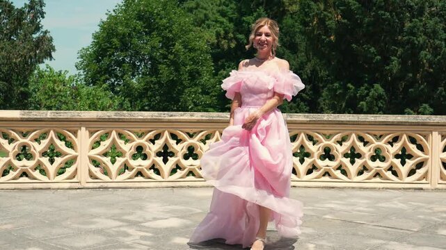 Lovely elegant woman in a pink ruffled wedding guest dress smiling and walking forward on a sunny architectural terrace.