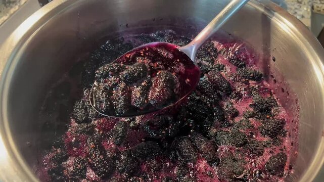 Checking the consistency of organic black mulberry jam with a spoon as it simmers in a pot. Details on traditional homemade flavors and dessert recipes.