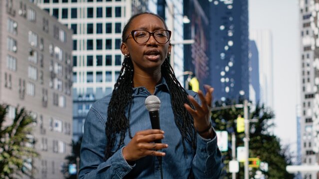 Presenting information on mic, field reporter girl downtown, on city street speaking towards camera, black journalist presenting breaking news reportage with updates, live commentary.