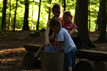 Fototapeta premium Mother spending quality time with her two young children sitting on a wooden bench in a peaceful forest. Warm sunlight filters through the trees, creating a cozy and intimate atmosphere. 