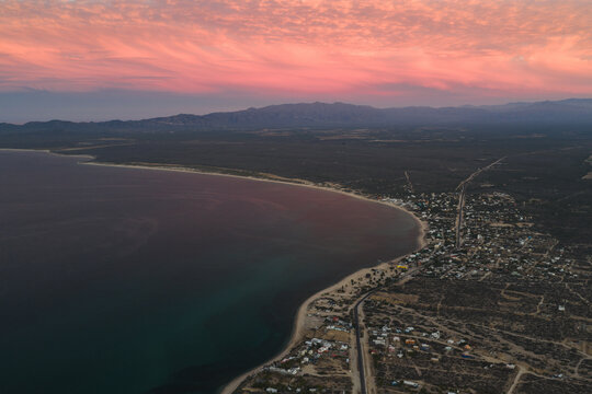 Aerial view of the coastal town of La Ventana at sunset, featuring the calm sea, sandy beach, and arid landscape under a vibrant pink sky in La Ventana, Baja California Sur, Mexico.