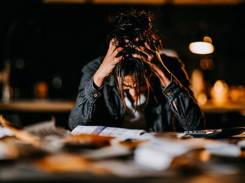 person sitting at a messy kitchen table hands rubbing their forehead in stress. Bills and papers spread out in front of them, a calculator nearby. . Expression of worry and exhaustion